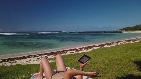 Eine-Aufnahme-Einer-Frau,-Die-Ein-Ebook-Auf-Dem-Gras-Eines-Tropischen-Strandes-Liest