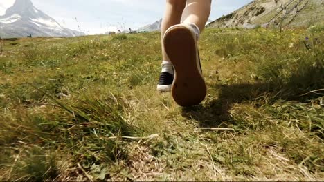 Low-dolly-shot-of-a-child's-feet-walking-on-grass-in-mountain-landscape-near-Zermatt,-Switzerland,-Matterhorn-partly-visible-in-background