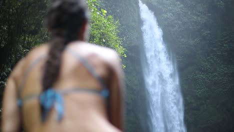 Toma-Panorámica-En-Cámara-Lenta-De-Una-Chica-En-Bikini-Azul-Sentada-Frente-A-Una-Cascada-De-Nungnung-En-Bali,-Indonesia