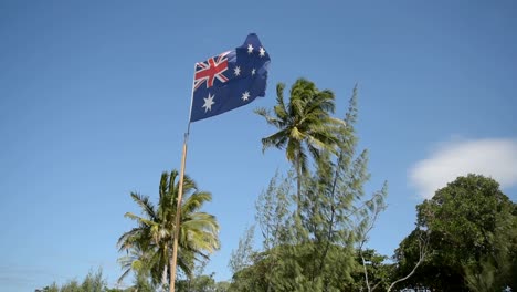 Hermosa-Bandera-Australiana-En-Cámara-Lenta-Con-Viento-Y-Hermosa-Palmera-Detrás-En-La-Playa-En-Verano
