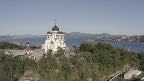 Slow-descent-aerial-shot-of-an-orthodox-church-with-blue-roof-and-golden-domes-with-port-and-bay-in-the-background-on-a-bright,-sunny-day