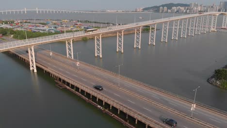 Drone-tilt-reveal-shot-of-Praia-Grande-bridge-passing-over-road-beside-Nam-Van-Lake,-with-Taipa-and-Outer-Harbour-in-background
