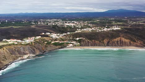 Praia-da-Arrifana-beach-in-west-Portugal-with-the-town-of-Picao-on-top-of-terrain,-Aerial-pan-right-shot