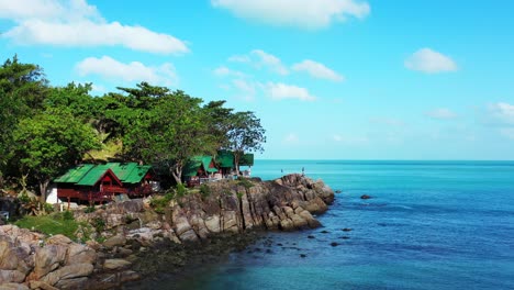 Beach-cabins-over-rocky-coastline-of-tropical-island-with-green-vegetation,-blue-turquoise-lagoon-reflecting-bright-sky-with-white-clouds-in-Vietnam