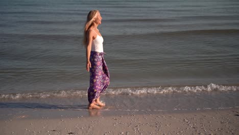 Slow-motion-of-mature-woman-in-sarong-walking-on-a-beach-and-throwing-her-arms-out-to-the-side-celebrating-freedom,-at-sunrise-or-sunset