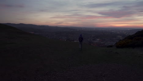 Cirlcing-parallax-shot-of-girl-taking-a-photo-with-smartphone-from-the-Arthurs-seat-mountain-in-evening,-dusk-with-city-of-Edinburgh-lit-up-in-the-background-during-wonderul-blue-hour