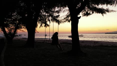 Silhouette-Eines-Mädchens,-Das-Auf-Der-Schaukel-Zwischen-Bäumen-Am-Tropischen-Sandstrand-Schwingt-Und-Einen-Friedlichen-Violetten-Sonnenuntergang-Genießt
