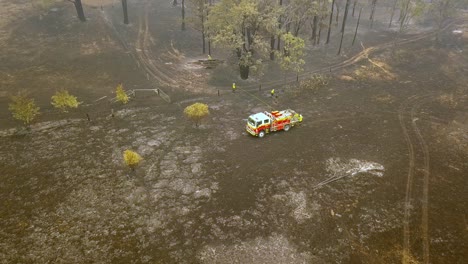Firefighters-inspect-the-scorched-burnt-ground-near-fire-engine-from-eucalyptus-forest-fire,-Aerial-drone-flyover-reveal-shot