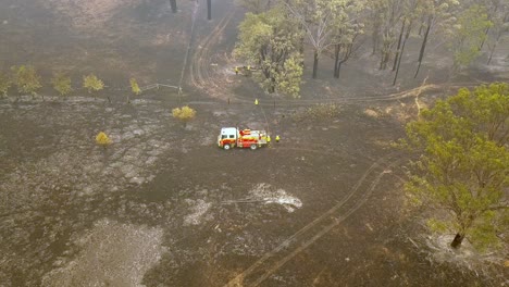 Firefighters-extinguish-embers-on-burning-ground-with-fire-engine-from-a-eucalyptus-forest-fire,-Aerial-drone-flyover-reveal-shot