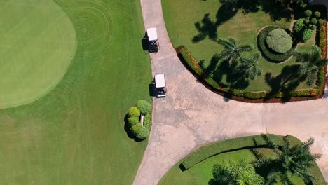 Golf-buggies-driving-along-path-of-lush-green-golf-course,-aerial-birds-eye-view