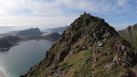 A-few-hikers-are-walking-up-the-trail-to-the-peak-of-mountain-Mannen,-overlooking-Haukland-beach-and-the-fjords-in-the-background-in-Lofoten,-Norway