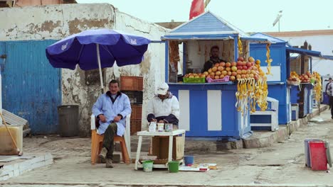 4k-Uhd-Szene-Von-Zwei-Alten-Fischern,-Die-Unter-Einem-Regenschirm-Im-Hafengebiet-Der-Alten-Marokkanischen-Stadt-Essaouira-Sitzen,-Mit-Einem-Obststand-Im-Hintergrund-Und-Einer-Vorbeilaufenden-Katze