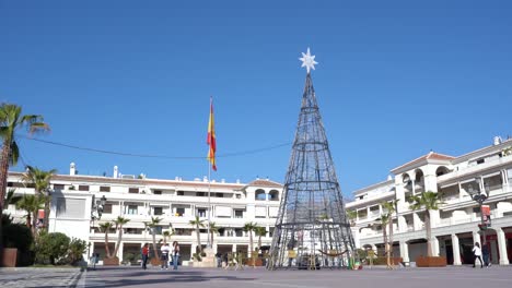 Tall-Christmas-Tree-And-Spanish-Flag-At-The-Plaza-De-Espana-In-Nerja,-Malaga,-Spain---ground-level-shot