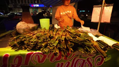 Mujer-Cocinando-Comida-Tradicional-Tailandesa-En-Hojas-De-Plátano-En-Un-Mercado-Nocturno
