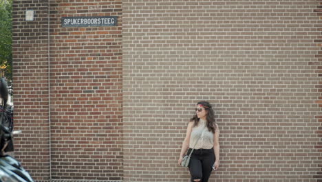 Stylish-Woman-with-Sunglasses-Leaning-and-Posing-in-front-of-Classical-Red-Brick-Stone-Wall-in-Cozy-Town-in-Netherlands-in-slowmo