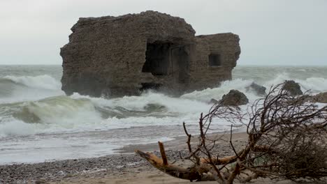 Big-stormy-waves-breaking-against-abandoned-seaside-fortification-building-ruins-at-Karosta-Northern-Forts-in-Liepaja,-Baltic-sea,-wave-splash,-pine-branches-in-front,-overcast-day,-medium-shot