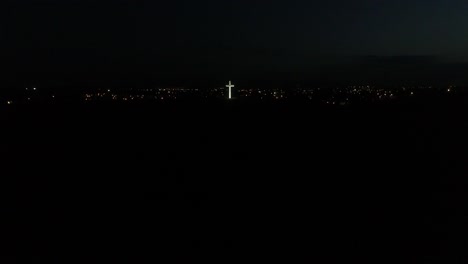 Breite-Aufnahme-Eines-Beleuchteten-Religiösen-Kreuzes-Auf-Einem-Hügel-Bei-Nacht-Mit-Stadtlichtern-Im-Hintergrund
