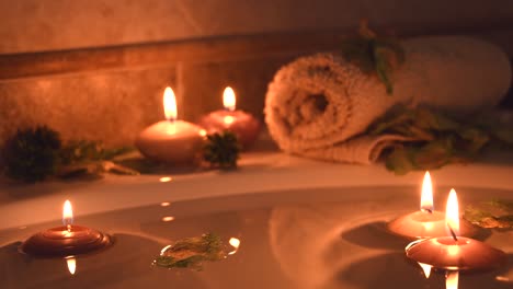 Entspannender-Spa-hintergrund-Mit-Im-Badewasser-Schwimmenden-Kerzen,-Einigen-Grünen-Blütenblättern-Und-Einem-Handtuch-In-Der-Nähe-Der-Wasseroberfläche