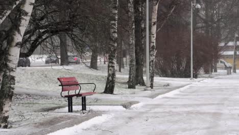 empty-bench-next-to-an-icy-road-in-a-park-during-a-snowstorm,-with-cars-in-the-distance