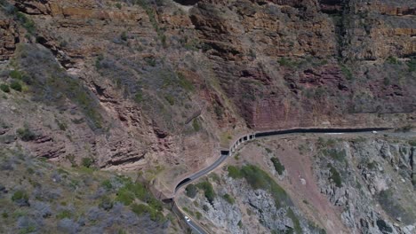 Drone-panning-shot-of-car-driving-on-scenic-coastal-road-cut-out-of-cliff-next-to-ocean---Chapman's-Peak-Drive,-Cape-Peninsula,-South-Africa