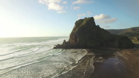 Aerial-wide-angle-of-Lion-Rock-Piha-tracking-towards-as-the-waves-gentle-crash-on-the-shore-line,-panning-into-the-sunlight-with-a-lens-flare