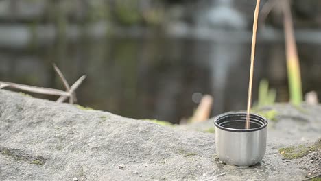 Pouring-hot-drink-from-flask-near-waterfall