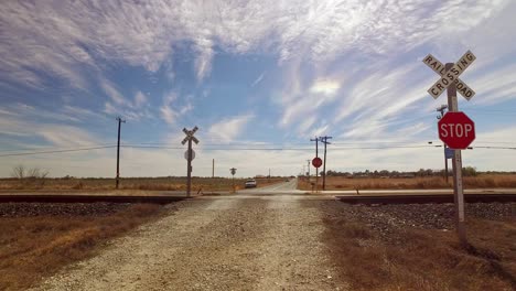 Car-approaching-a-railroad-crossing-out-in-the-middle-of-nowhere,-long-road-with-wide-open-fields-and-blue-sunny-skies