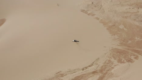 Aerial:-A-truck-with-kitesurfers-traveling-through-the-dunes-of-Lencois-Maranhenses-in-Brazil,-during-dry-season