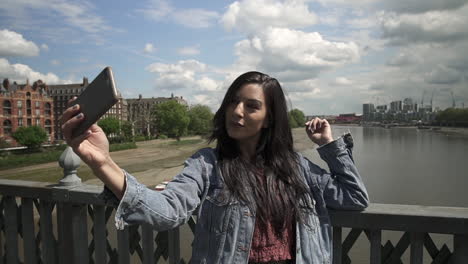 Brunette-Latina-tourist-taking-a-selfie,-posing-doing-victory-sign-with-her-hand,-while-standing-on-the-railing-of-a-bridge-in-London