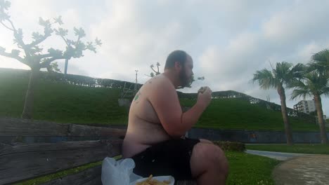 Shirtless-man-eating-a-burger-while-exposing-his-fat,-ugly-stomach-sitting-on-public-park-bench
