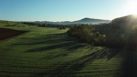 aerial-drone-view-of-morning-hour-over-peaceful-countryside-with-green-and-agricultural-fields,-sun-shining-directly-into-camera,-switzerland