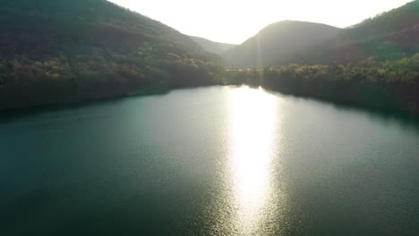 Toma-Aérea-Cinematográfica-Sobre-El-Lago-Del-Bosque-Rodeado-De-Montañas-Verdes-Al-Atardecer