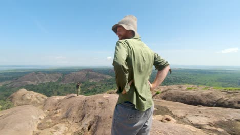 Slow-motion-orbiting-shot-around-a-bearded-ginger-man-with-a-large-hat-while-standing-on-top-of-a-granite-mountain-and-looking-out-to-East-African-views