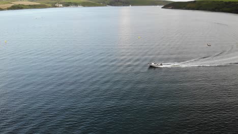 Drohnenaufnahmen-Eines-Motorboots,-Das-Auf-Dem-Wasser-Mit-Fernen-Grünen-Hügeln-Und-Segelbooten-Rast