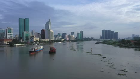 drone-shot-of-A-cargo-ship-and-smaller-vessel-transporting-shipping-containers-along-the-Saigon-river-with-the-financial-and-city-center-in-full-view