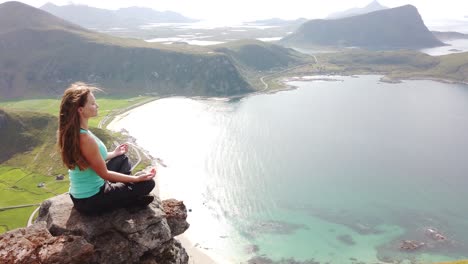 A-young,-white-female-hiker-is-sitting-down-and-meditating-on-a-rock-on-the-edge-of-a-cliff-on-top-of-Mannen-mountain,-overlooking-Haukland-beach-during-a-warm-and-sunny-summer-day-in-Lofoten,-Norway