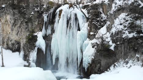 Aufsteigender-Schuss-über-Teilweise-Gefrorenem-Wasserfall,-Um-Berge-Und-Wald-Im-Winter-Mit-Schnee-In-4k-Zu-Enthüllen