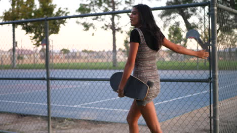 A-pretty-young-hispanic-woman-smiling-and-walking-through-an-urban-city-park-with-her-skateboard-along-a-chainlink-fence-SLOW-MOTION