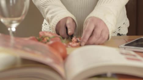 Manos-Cortando-Tomates-De-Vid-Frescos-En-La-Cocina-Con-Libro-De-Recetas-En-Primer-Plano