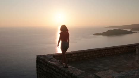 Beautiful-lady-walking-on-a-viewpoint-looking-over-the-Montenegro-coastline-over-top-of-Sveti-Stefan-during-a-beautiful-Sunset