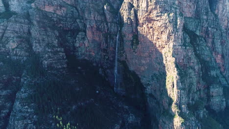 Aerial---Ascending-drone-with-camera-tilting-down-revealing-waterfall-falling-down-imposing-cliff-face-of-sandstone-mountain