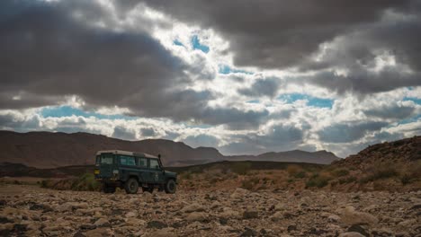 Time-lapse-of-clouds-and-sun-rays-over-4WD-vehicle-parked-in-desert,-wide-angle