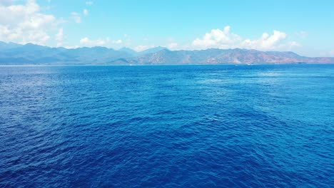 El-Mar-Azul-Ondulado-Alrededor-De-La-Isla-De-La-Montaña-Se-Ilumina-Con-La-Luz-Del-Sol-De-La-Mañana-En-Lombok,-Indonesia,-Panorama-Aéreo-Con-Espacio-Para-Copiar