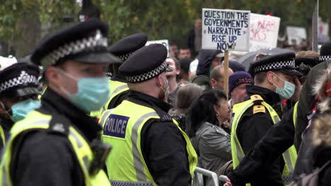 A-line-of-police-in-protective-face-masks-stand-watching-a-Coronavirus-and-QAnon-conspiracy-protest,-as-one-protestor-holds-a-placards-that-says,-“Question-the-government’s-narrative,-rise-up-now”