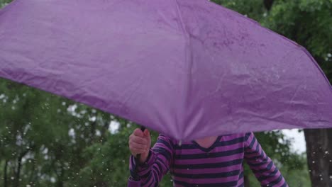 Close-up-view-of-young-Caucasian-girl-wearing-striped-shirt-holding-purple-umbrella-jumps-up-and-down-outside-splashing-water-in-puddle,-static-portrait-slow-motion