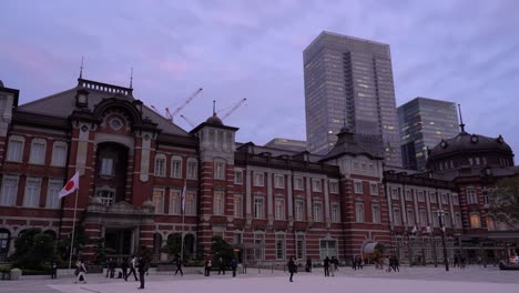 Locked-off-sideways-view-out-on-Tokyo-station-with-skyscrapers-in-background-and-people-wearing-facemasks-walking-through-the-frame