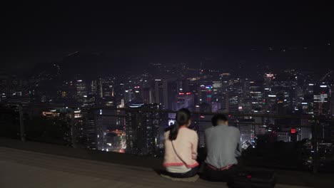 A-couple-wearing-medical-masks-having-a-date-at-Namsan-tower-watching-Night-Seoul,-focuses-on-city-buildings,-a-couple-is-not-in-focus