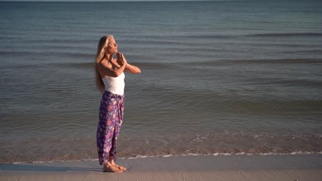 Slow-motion-of-mature-woman-in-sarong-in-a-prayer-pose-on-a-beach-at-sunrise-or-sunset