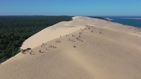 People-walk-on-top-of-Dune-du-Pilat-in-the-Arcachon-Bassin-with-a-height-of-over-100-meters,-Aerial-flyover-shot