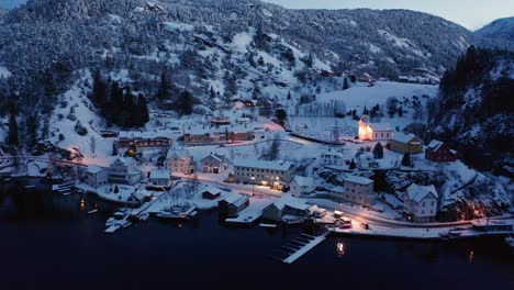 Aerial-of-small-Idyllic-Norwegian-village-Stamnes-at-dusk-during-wintertime---Street-lights-and-church-in-frozen-landscape